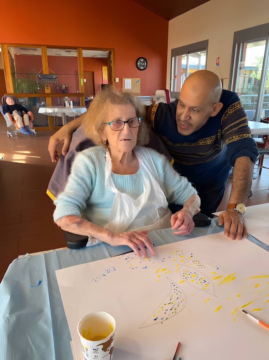 Une femme âgée aux cheveux gris clairs et fins, portant des lunettes et un pull bleu clair, est assise à une table et peint sur une grande feuille de papier blanc. Un homme au crâne chauve et portant un pull rayé se tient debout à côté d'elle, penché vers elle, une main sur son épaule, souriant.